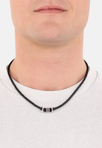 Schwarzes geflochtenes Lederhalsband mit einem zylindrischen Metallelement in Silber und Schwarz, getragen zu einem weißen Rundhalsausschnitt-Shirt.