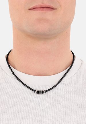 Collana di pelle intrecciata nera con un accento metallico cilindrico con strisce nere e argento, indossata su una maglietta bianca a girocollo.