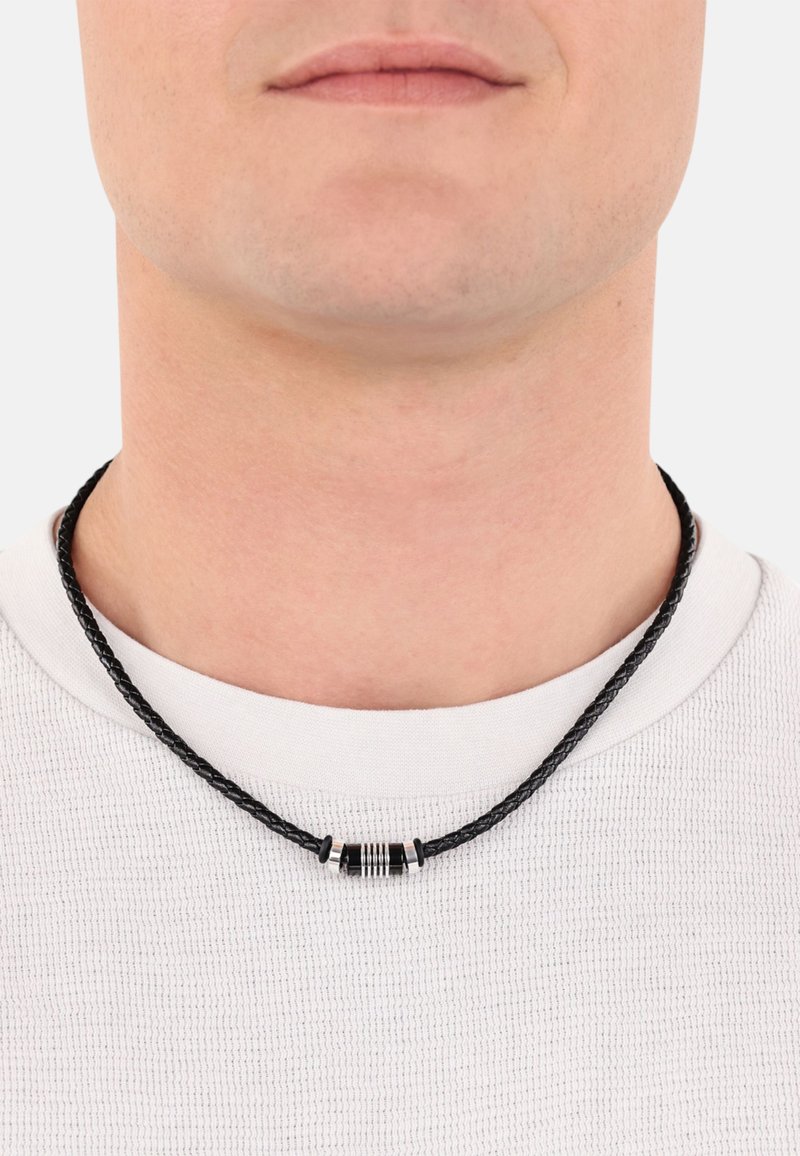 Schwarzes geflochtenes Lederhalsband mit einem zylindrischen Metallelement in Silber und Schwarz, getragen zu einem weißen Rundhalsausschnitt-Shirt.