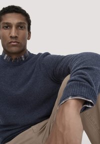 Navyblauer Wollpullover mit gerippten Bündchen über einem karierter Hemd. Hellbraune Hose mit Seitentaschen, sitzt vor einem neutralen Hintergrund.