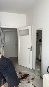 Balta patalpa su medinėmis durimis su stiklo elementu, pilno ilgio veidrodžiu su plonu rėmu ir šviesios spalvos grindų kilimu su rudais akcentais.