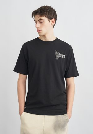 Jeune homme aux cheveux courts et foncés portant un T-shirt noir avec le texte "volcon stone" et un graphisme sur la poitrine, les mains dans les poches d'un pantalon beige.