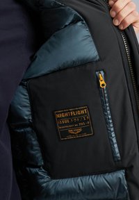 Zwarte puffer vest met een marineblauwe voering, voorzien van een voorste patchlabel, een ritsvak en een oranje ritsenhanger. Getextureerde stof en glanzende accenten.