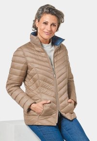 Giacca trapuntata beige chiaro con colletto alto e fodera interna blu. Presenta una chiusura con zip e tasche laterali. Indossata sopra un top bianco.