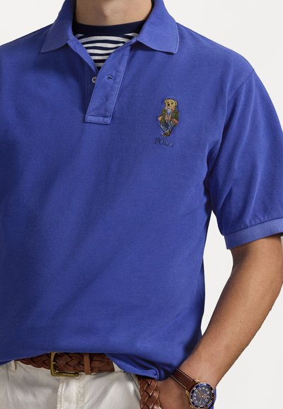 Královsky modré polo tričko s žebrovaným límcem, na kterém je vyšitý medvěd v saku a pod ním slovo "POLO". Nosené s hnědým pleteným opaskem.