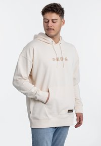 Beige hoodie met trekkoorden en een buidelzak. Heeft subtiele grafische ontwerpen in lichtbruin boven de zak. Zachte textuur.