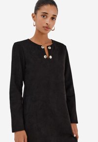Černé semišové šaty s dlouhými rukávy, kulatým výstřihem a třemi ozdobnými stříbrnými knoflíky na krku. Hladká textura a přiléhavý střih.