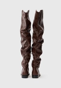 Botas de cuero marrón, hasta la rodilla, con un diseño holgado y un acabado texturizado y un ligero tacón. La parte trasera presenta pliegues gatherados y un acento de cuero liso.