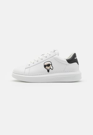 Baskets en cuir blanc avec un accent noir au talon. Présente une patch cartoon d'une figure avec des lunettes de soleil. Bout arrondi et épaisse semelle blanche.