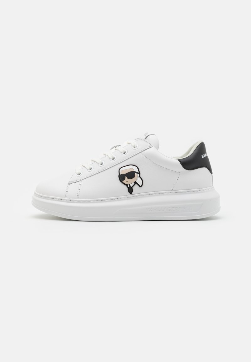 Sneaker bianca in pelle con accento nero sul tallone. Presenta una toppa cartoon di una figura con occhiali da sole. Punta arrotondata e suola spessa bianca.