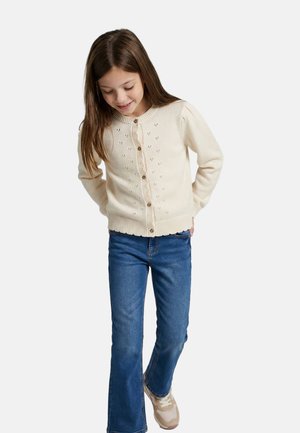 Jeune fille aux longs cheveux bruns portant un cardigan crème boutonné, un jean bleu et des baskets beiges, regardant vers le bas et souriant.