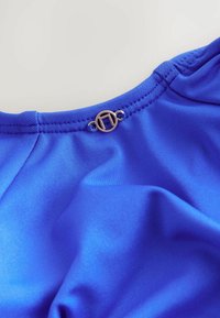 Tissu bleu royal au toucher lisse, avec un fermoir en métal circulaire au niveau du col. Des coutures sont visibles le long des bords.