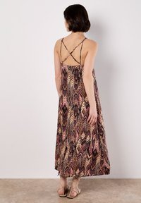 Un modello sta di spalle rispetto alla fotocamera, indossando un maxi dress stampato con spalline sottili e sandali con cinturini, su uno sfondo neutro.