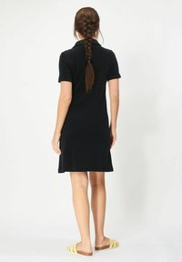 Svart poloklänning med korta ärmar, i midjehöjd, med sidslitsar. Bärs med beige slip-in sandaler, håret i flätad stil.