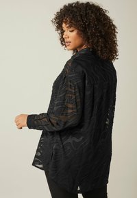 Black sheer blouse with textured wavy pattern, long sleeves, buttoned cuffs, and a relaxed fit, featuring a rounded hem.