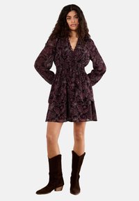Robe à manches longues, imprimé paisley sombre, avec une taille cintrée, une jupe à volants et un décolleté en V profond. Associée à des bottes en daim marron jusqu'au genou.