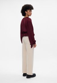 Blusa pregueada bordeaux com mangas longas, atada à cintura, combinada com calças largas creme e botas de plataforma pretas.