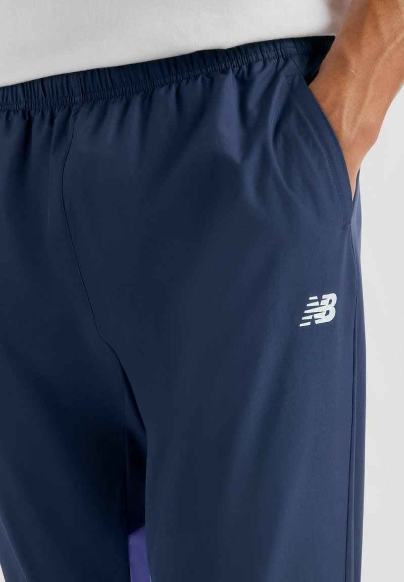 Person wearing navy blue athletic shorts with elastic waistband and white New Balance logo, hand placed inside the pocket.