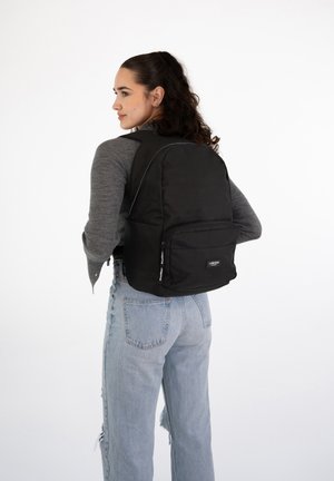 Schwarzer Stoffrucksack mit gepolsterten Trägern, einer Vorderfronttasche und weißen Nähdetails, getragen auf dem Rücken einer Person in hellblauen Jeans.