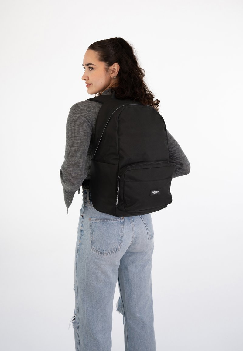 Black fabric backpack with padded straps, a front pocket, and white stitching details, worn on the back of a person in light blue jeans.