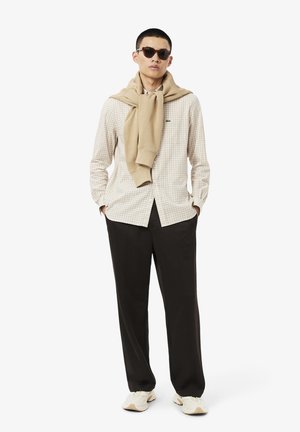 Camicia a quadri di colore chiaro, maglione beige appoggiato sulle spalle, pantaloni neri e scarpe da ginnastica chiare. Il modello indossa occhiali da sole scuri.