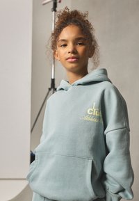 Vaaleansininen hupullinen collegepaita, tehty pehmeästä kankaasta, jossa on etutasku ja keltainen logo. Rentoutunut istuvuus resorireunuksilla.