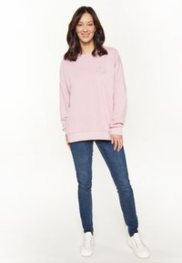 Felpa rosa con maniche lunghe e polsini a coste, con un delicato design floreale sul petto, abbinata a jeans blu e sneakers bianche.