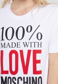 Bílý bavlněný tričko s výrazným černým a červeným textem, který říká "100% VYROBENÉ S LÁSKOU MOSCHINO" v různých typech písem. Krátké rukávy.