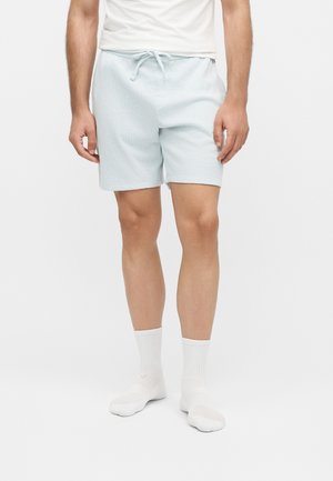 Mann trägt hellblaue Shorts mit Kordelzug, weiße Sportsocken und ein weißes T-Shirt und steht vor einem schlichten weißen Hintergrund.