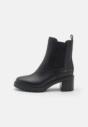 Botte de cheville en cuir noir avec un talon épais, panneau latéral élastique, tirant à l'arrière et petit logo doré Tommy Hilfiger sur le côté.