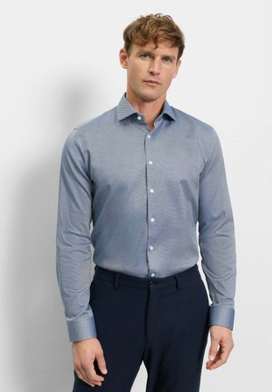 Mann trägt ein hellblaues, langärmeliges Hemd mit Muster und dunkle blaue Hosen, steht vor einer schlichten weißen Wand.