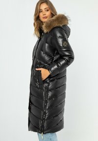 Schwarze Daunenjacke mit Kapuze und Kunstfellbesatz, mit einem gesteppten Design, Reißverschlusstaschen und einem goldfarbenen Logo-Akzent am Ärmel.