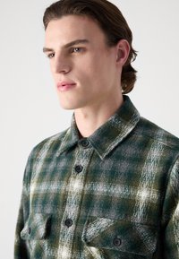 Green and gray plaid flannel shirt with two chest pockets, black buttons, and a point collar, featuring a soft, textured fabric finish.