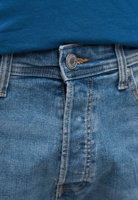 Κοντινό πλάνο της μέσης ενός μπλε τζιν με μεταλλικό κουμπί κλεισμένο και μέρος μιας μπλε πουκάμισου που έχει μπει μέσα στο παντελόνι να φαίνεται από πάνω.