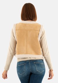 Gilet en daim beige avec une finition en fausse fourrure, présentant une coupe décontractée, des emmanchures ouvertes et une texture lisse, associé à un jean bleu.