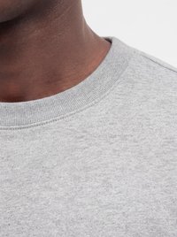 Close-up af en persons nakke og øverste bryst, der bærer en lysegrå crewneck sweatshirt mod en ensfarvet baggrund.