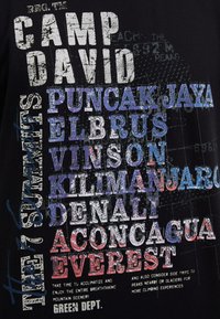 Black t-shirt featuring multicolored text: "CAMP DAVID," mountain names, and additional text. The design incorporates a textured print effect.