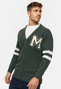 Groene gebreide cardigan met kabelpatroon, V-hals, knoopsluiting en een geborduurde 'M' op de borst; beschikt over witte gestreepte accenten op de mouwen.