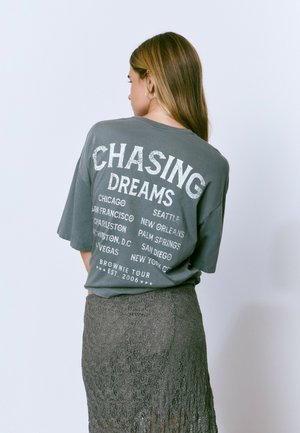 T-shirt gris surdimensionné avec un texte graphique blanc "CHASING DREAMS" et des noms de villes au dos, associé à une jupe en dentelle texturée.