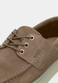 Chaussure en daim marron à bout arrondi, design classique à lacets, œillets argentés et semelle en caoutchouc beige. Présente un logo embossé sur le côté.