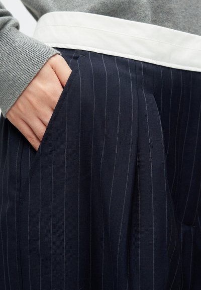 Jūras spēku vertikāli svītrainā svārku ar krokām un baltu jostasvietu. Audums izskatās teksturēts, un dizainā ir sānu kabatas.