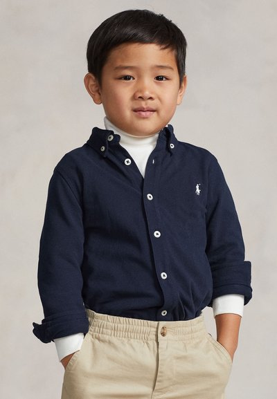 Jeune garçon les mains dans les poches, portant une chemise bleu marine boutonnée sur un col roulé blanc et un pantalon beige, debout devant un fond neutre.