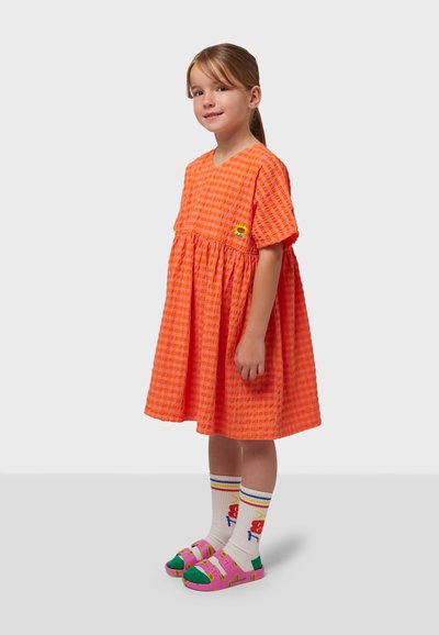 Orange-rutig klänning med korta ärmar och draperad midja. Kombinerad med rosa sandaler och mönstrade vita strumpor med färgglada accenter.