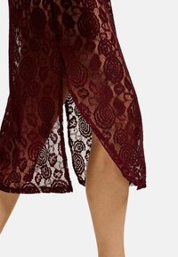 Burgundna čipkasta obleka z cvetličnim vzorcem, z razporkom na strani. Prosojna tekstura poudarja zapletene detajle in plastičen dizajn.