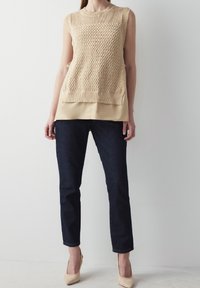 Beige stickad ärmlös topp över ett krämigt lager, parat med mörk denimjeans och nude spetsiga klackar. Mönstrad textur på toppen.