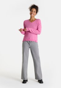 Rosa V-Ausschnitt-Pullover aus weichem Material, kombiniert mit grauen, weit geschnittenen Hosen. Der Pullover hat lange Ärmel und ein dezent seitliches Akzentdetail.