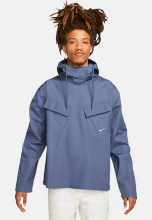 Giacca in nylon blu con cappuccio regolabile, due tasche frontali e logo Nike. Presenta una vestibilità ampia e una texture liscia.