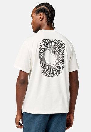Mand med fletninger iført en hvid t-shirt med et sort hvirvlende "Brooks Running" logo på ryggen, kombineret med blå bukser.