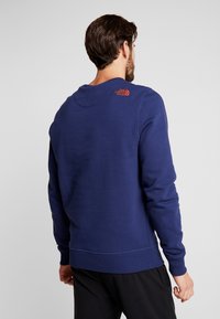 Man med en marinblå sweatshirt med The North Face-logotypen på övre ryggen, vänd bort från en enkel ljusgrå bakgrund.