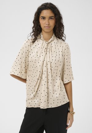 Vrouw met krullend haar, gekleed in een beige blouse met zwarte stippen en een strikdetail aan de voorkant, gecombineerd met een zwarte broek en een gouden armband.
